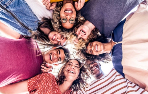 Multicultural group of young people standing in circle and smiling at camera - Happy diverse friends having fun hugging together - Low angle view